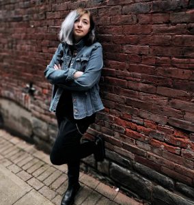 a young woman wearing denim and black leans against a brick wall