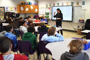 a woman wearing dark clothing stands in front of a smart board, teaching students in a classroom