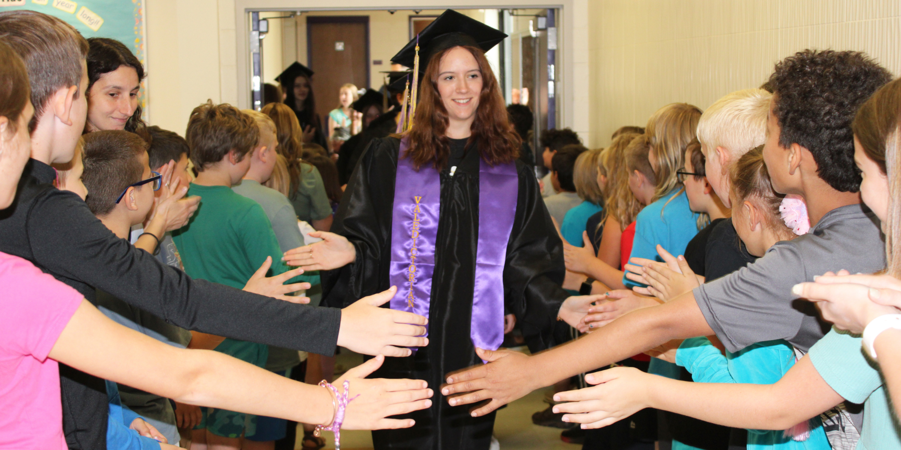 a young woman wearing a graduation gown walks through the hallway and high fives younger students