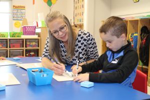 a woman sits at a blue table with a young student as they work on a project together