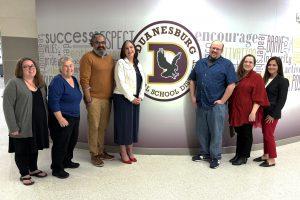 a group of five women and two men pose next to a school logo in the hallway