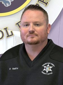 a man with facial hair wearing a black shirt poses in front of a purple wall with a logo on it