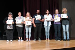 Male and female teachers pose on a stage holding certificates.