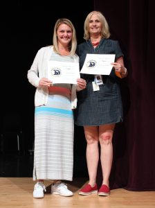 two blonde women hold certificates on a stage.