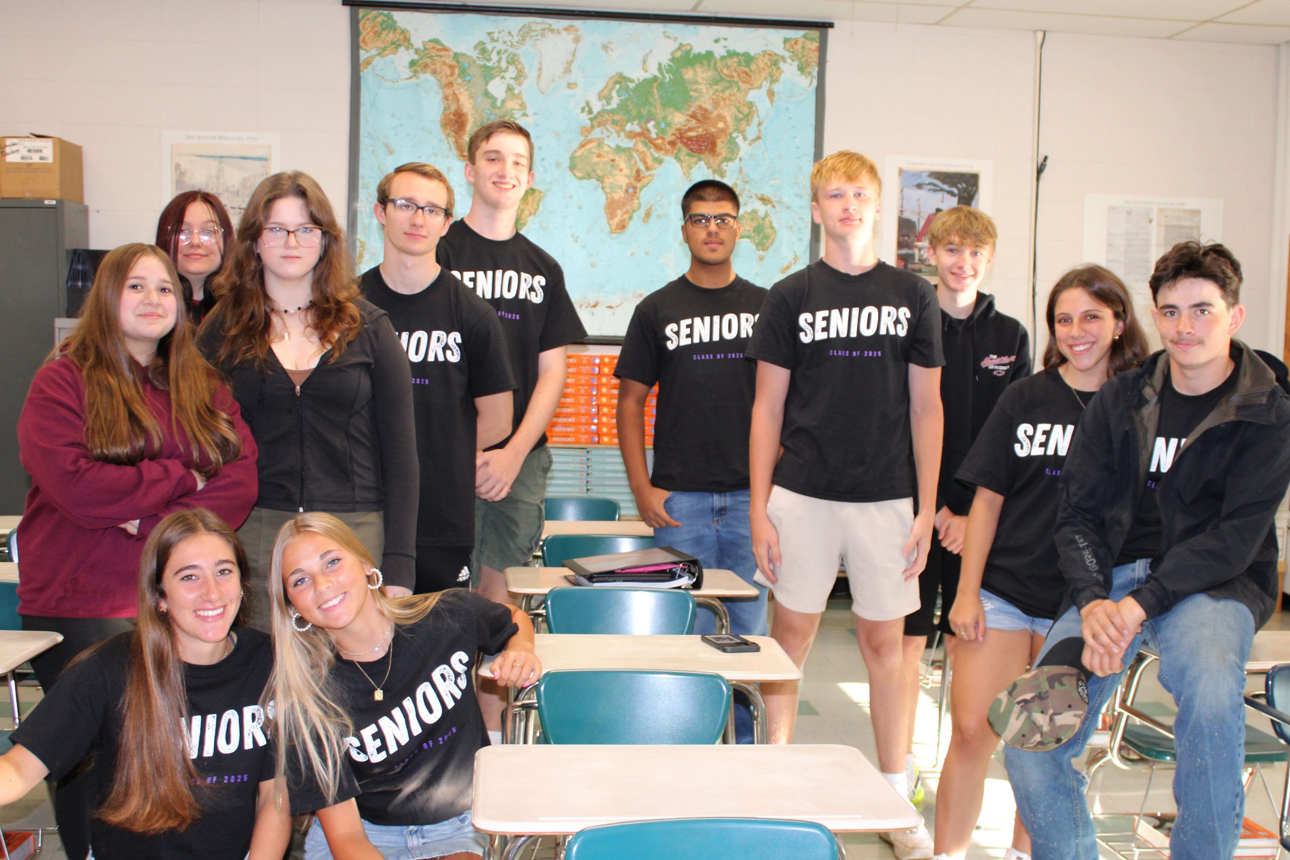 teenagers wear black shirts that say "SENIOR" on the front, while posing in a classroom