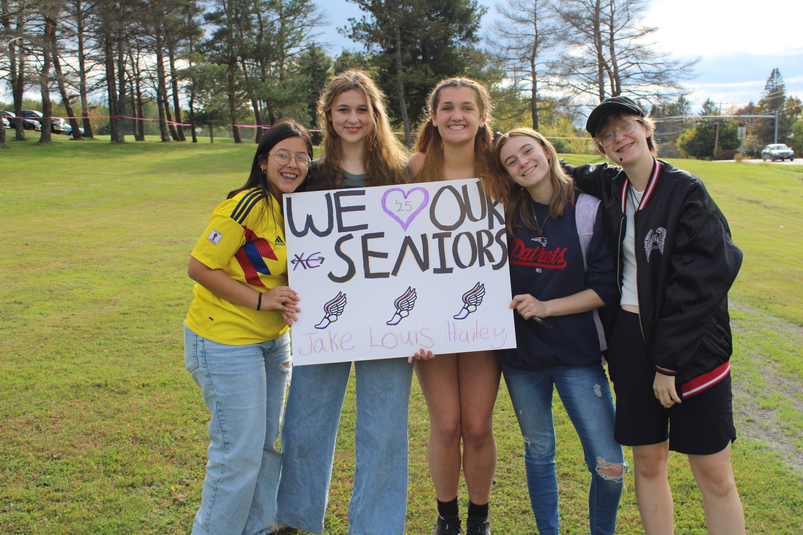 teenaged girls pose together holding a sign that reads "WE LOVE OUR SENIORS"