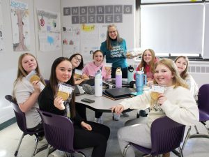 a woman poses with students sitting at a table, holding up cookies