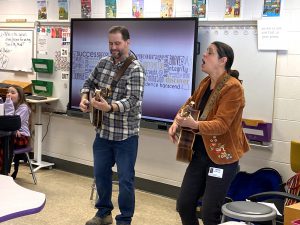 a man and a woman play guitar in front of a classroom of students